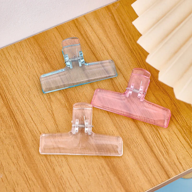 Drei klare und rosa Acryl-Ordnerclips sind auf einer Holzoberfläche arrangiert.