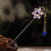 Une épingle à cheveux en bois foncé avec une fleur violette clair et des accents dorés, ornée de perles bleues.