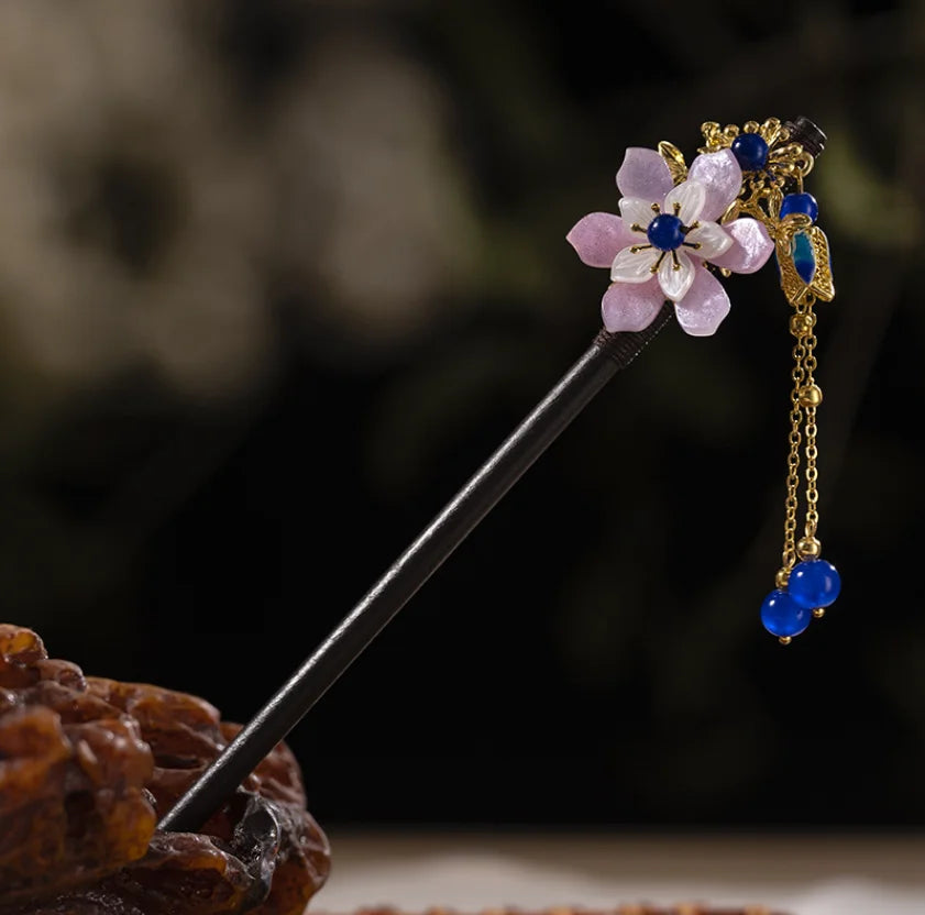 Une épingle à cheveux en bois foncé avec une fleur violette clair et des accents dorés, ornée de perles bleues.