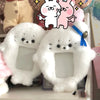 White plush photocard holder with a soft cover and blue ribbon, displayed next to pink plush characters.