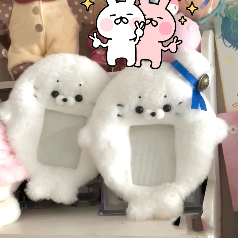 White plush photocard holder with a soft cover and blue ribbon, displayed next to pink plush characters.