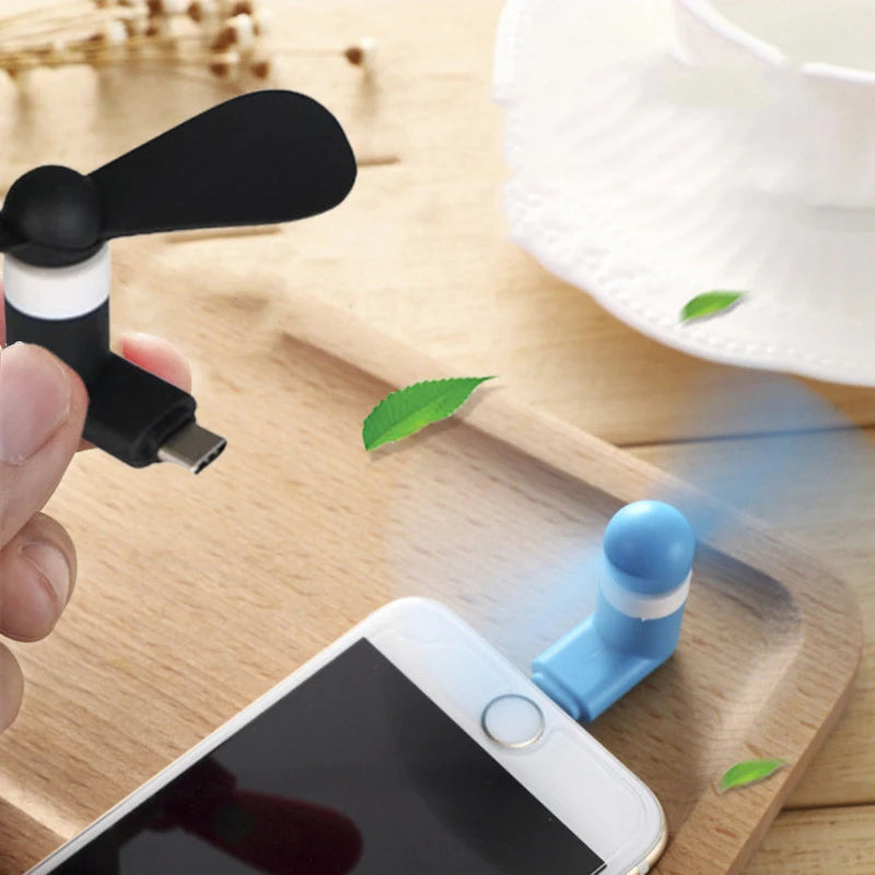 Una mano sostiene un ventilador USB portátil negro junto a un smartphone sobre una tabla de cortar de madera.
