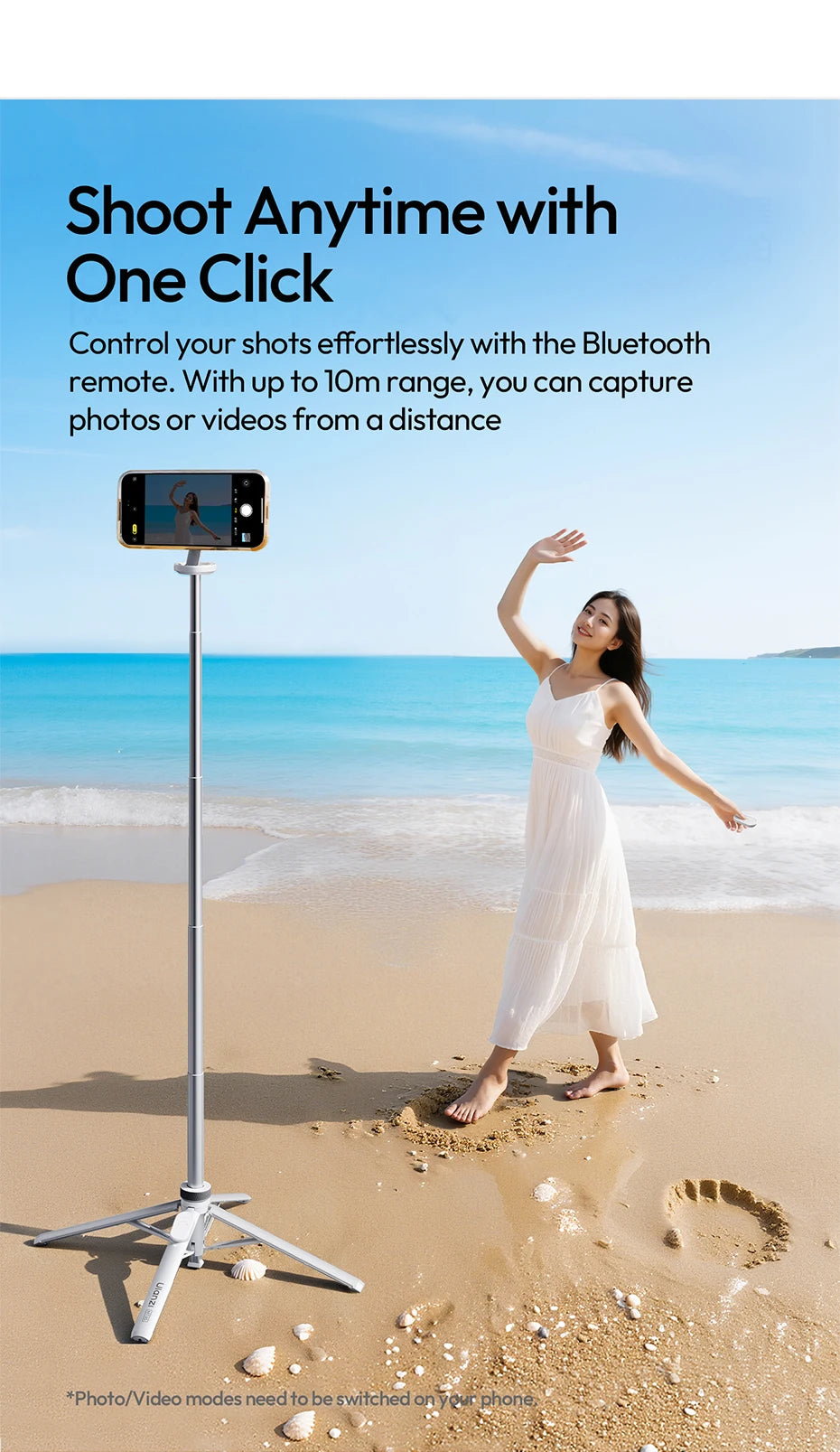 A woman stands on a beach holding a white magnetic phone tripod with a Bluetooth remote.