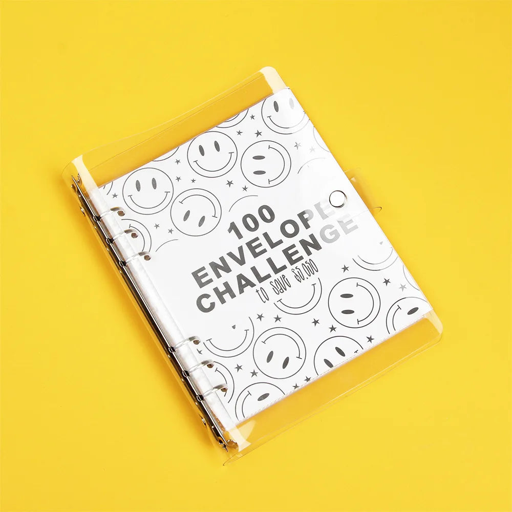 A clear plastic sleeve holds a spiral notebook titled 100 Envelope Challenge with smiley face icons on the cover.