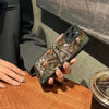 Une main tient une coque de téléphone en silicone camouflage de forêt pour iPhone sur une surface en bois.