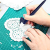 A hand uses a dark blue steel art cutting tool to cut intricate lace patterns into white paper on a green cutting mat.
