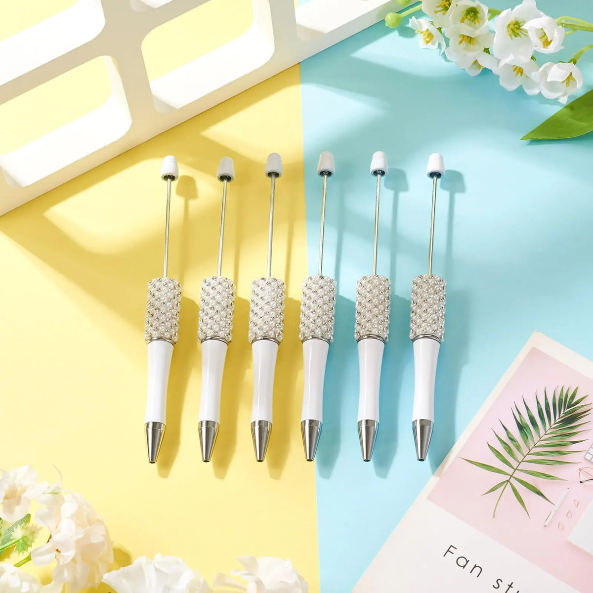 Five pearl beaded ballpoint pens displayed on a light blue and yellow background with white flowers.
