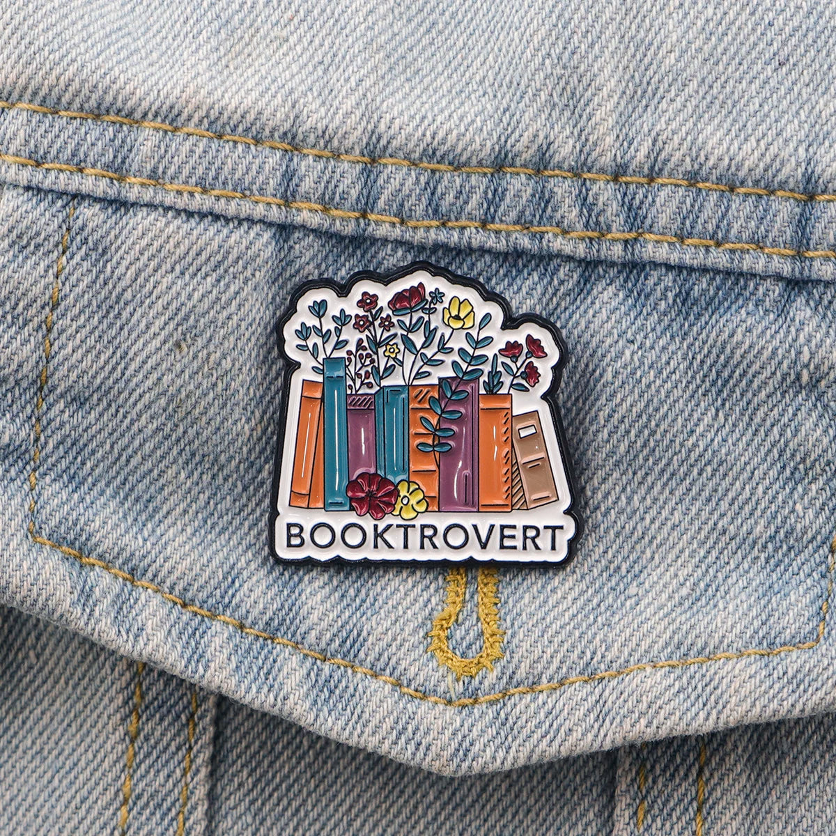 An enamel lapel pin featuring a stack of colorful books with floral accents and the word BOOKTROVERT.