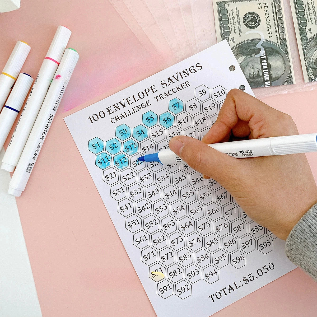 A hand writes on an envelope savings tracker sheet showing various dollar amounts and a total of $5,050.