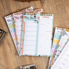 A stack of weekly meal planner and grocery list pads with pink and green covers are displayed on a wooden surface.