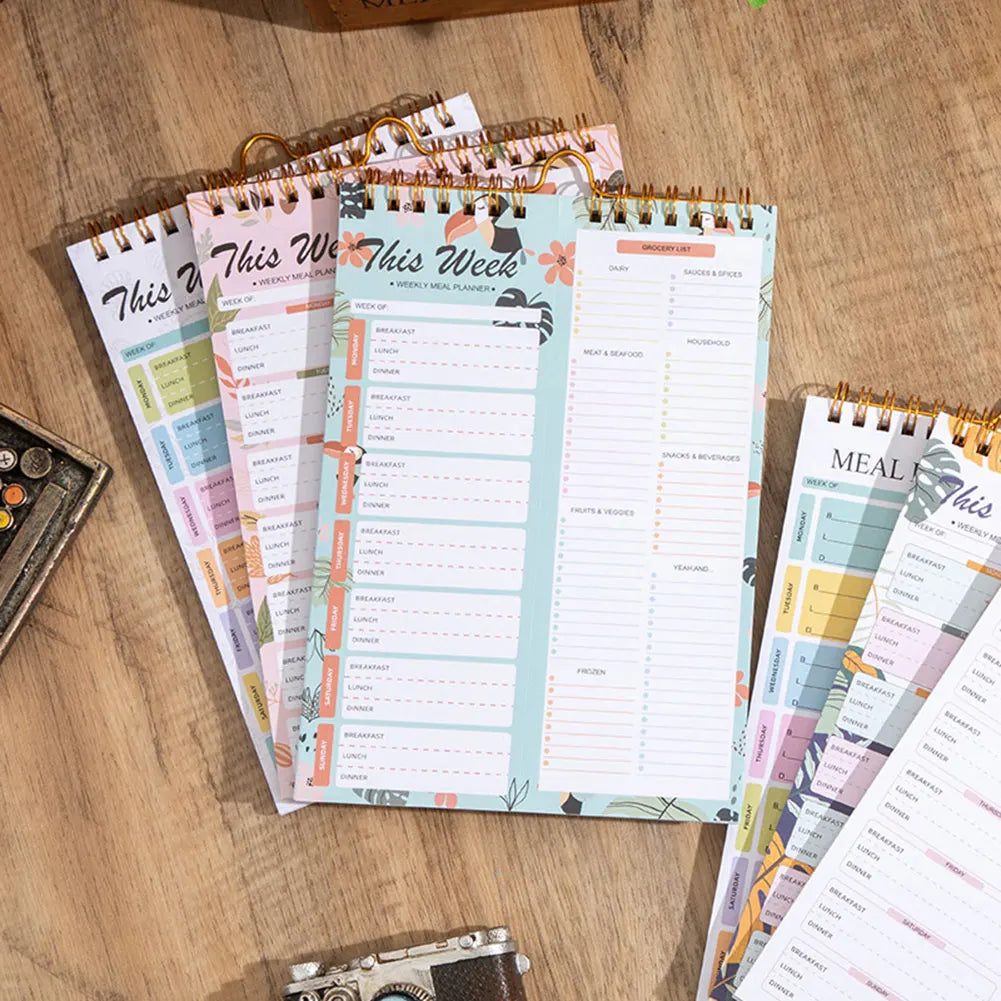 A stack of weekly meal planner and grocery list pads with pink and green covers are displayed on a wooden surface.