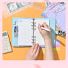 A hand is writing in a blue A6 binder with a grid layout on an orange background surrounded by financial documents.
