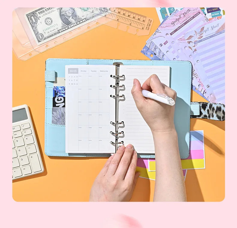 A hand is writing in a blue A6 binder with a grid layout on an orange background surrounded by financial documents.