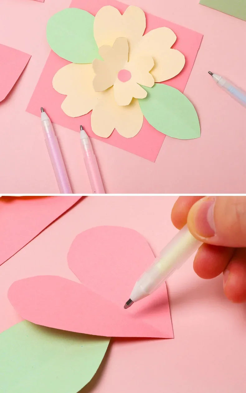 A hand is writing on a pink paper heart using a white glue pen, surrounded by various colored paper shapes.