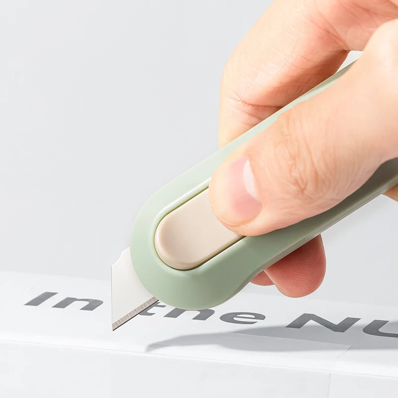 A hand is operating a light green compact mini paper cutter with a beige blade on a white box.