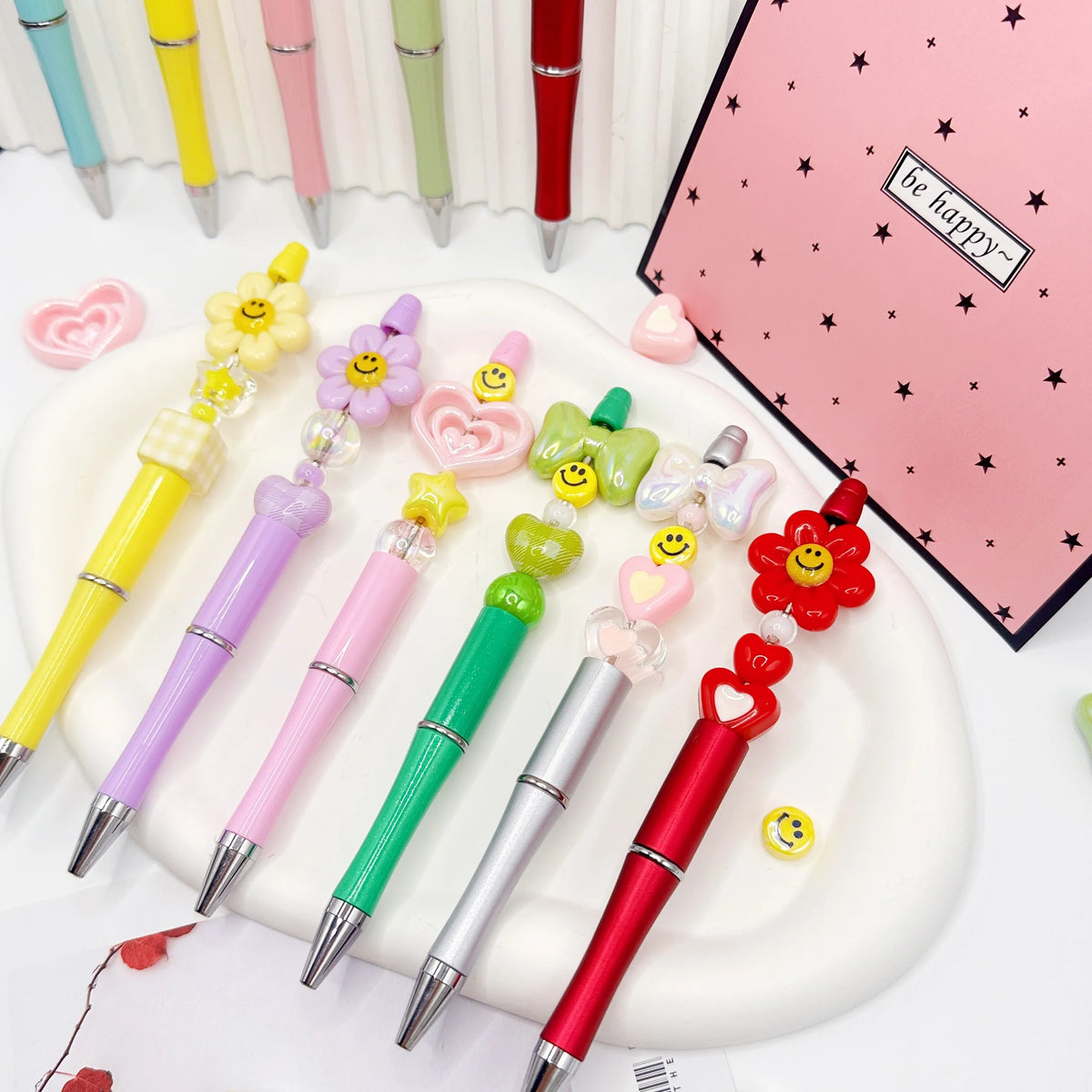 A set of colorful beaded ballpoint pens arranged on a white plate with a pink notebook.