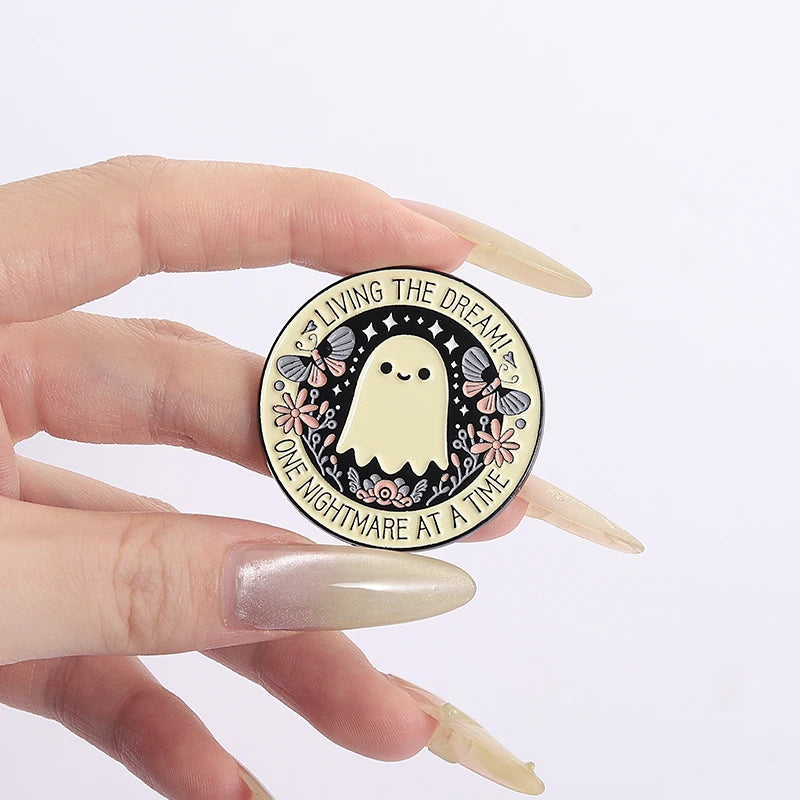 A hand holds a round enamel pin featuring a white ghost with butterfly wings and text that reads Living the Dream One Nightma