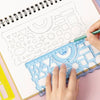 A hand is using a clear plastic ruler to draw geometric shapes on a spiral notebook page.