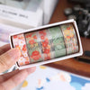 A hand holds a box of decorative transparent washi tape rolls featuring floral patterns in various colors.