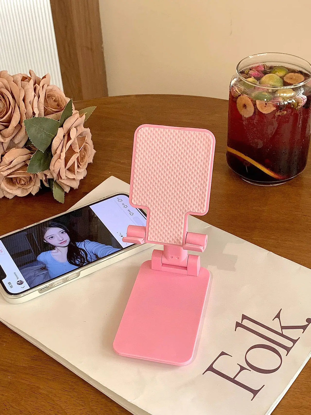 A pink adjustable foldable phone and tablet stand resting on a wooden table next to a glass of iced tea.