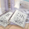 A clear plastic case holds a monthly savings challenge money clip featuring floral designs and text.
