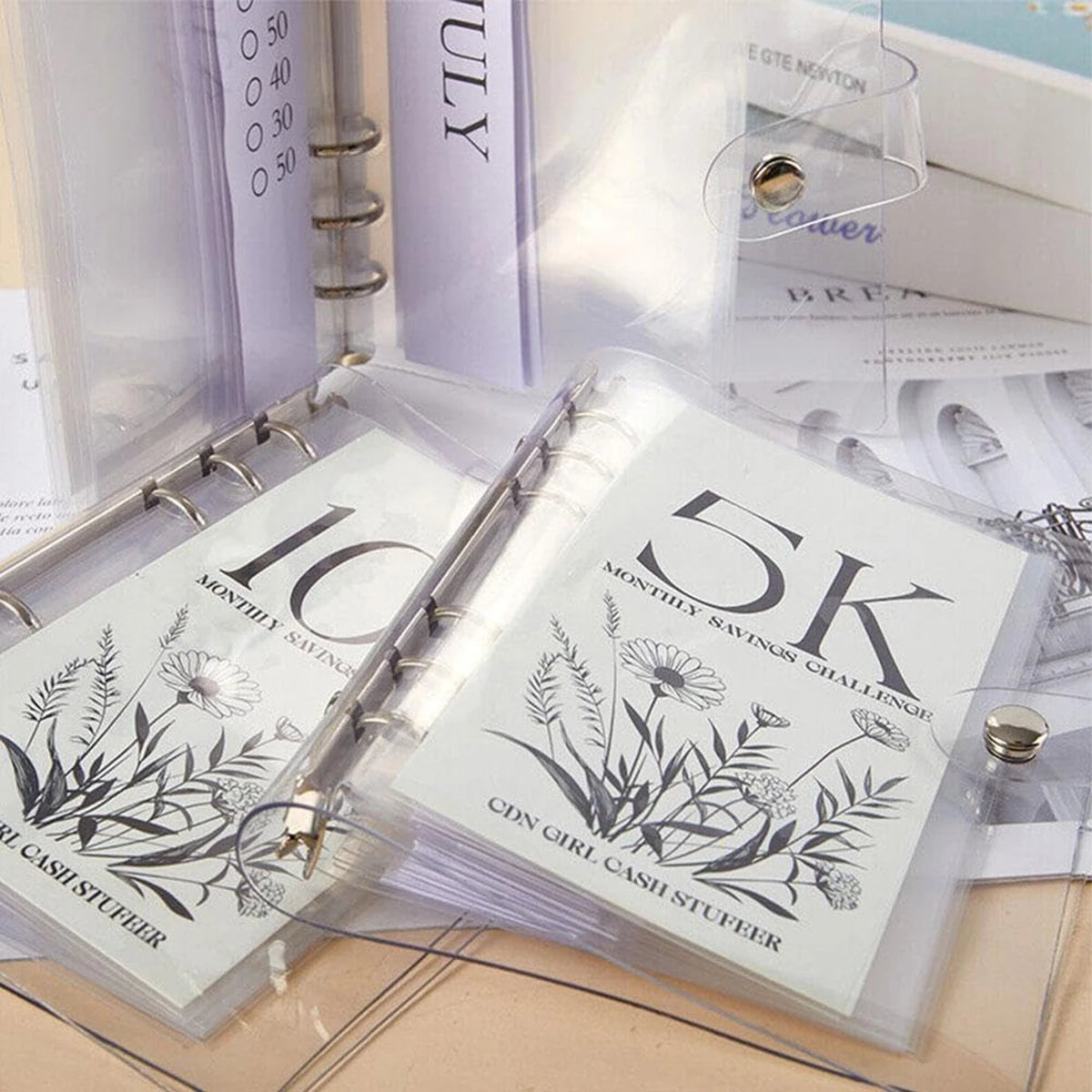 A clear plastic case holds a monthly savings challenge money clip featuring floral designs and text.