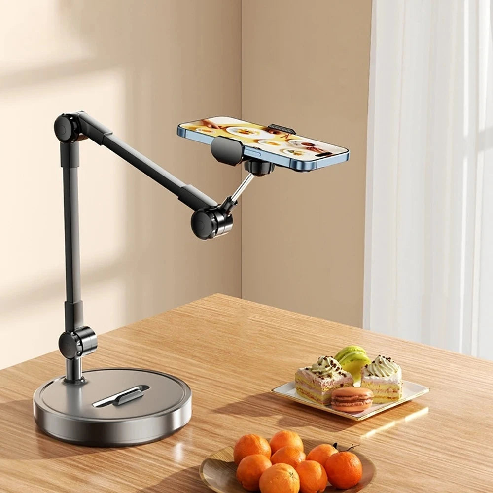 A black multi-angle adjustable phone holder stand is shown on a wooden table with fresh fruit nearby.