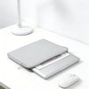 A silver laptop is placed inside a gray waterproof PU leather sleeve on a white desk next to a white lamp and mouse.