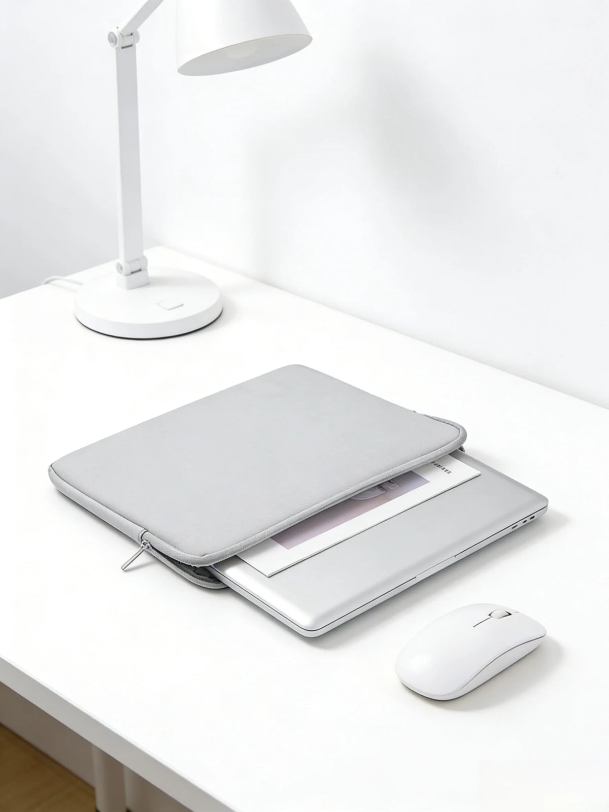 A silver laptop is placed inside a gray waterproof PU leather sleeve on a white desk next to a white lamp and mouse.