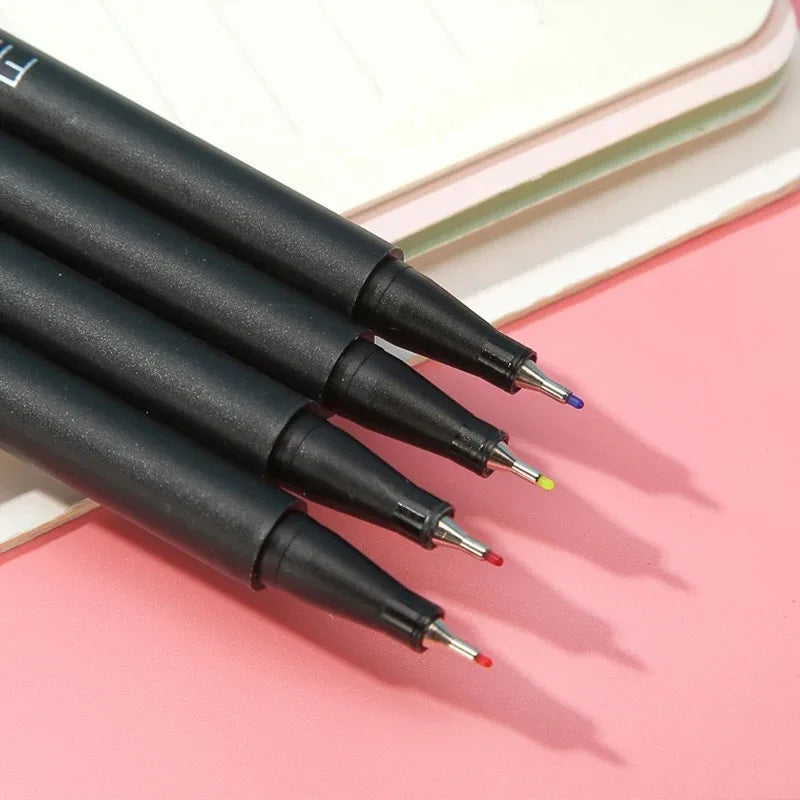 A set of black fine line marker pens with various colored tips resting on a pink surface.