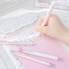 A hand holds a pink gel pen, with several pearl angel-themed pens and decorative cutouts on a pink notebook.