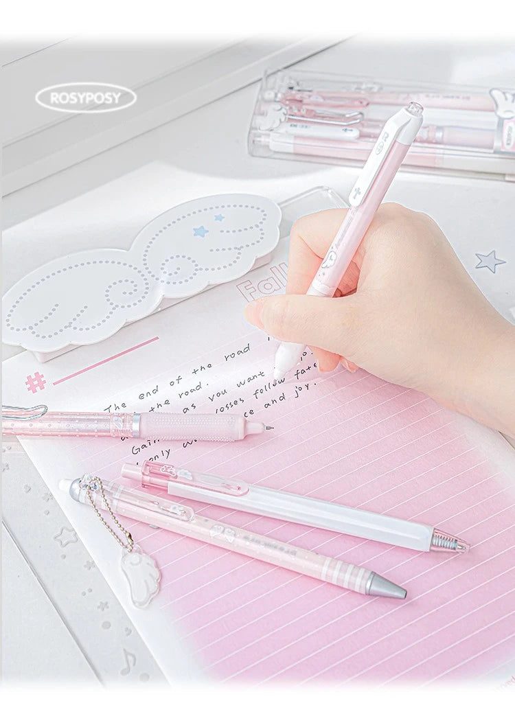 A hand holds a pink gel pen, with several pearl angel-themed pens and decorative cutouts on a pink notebook.