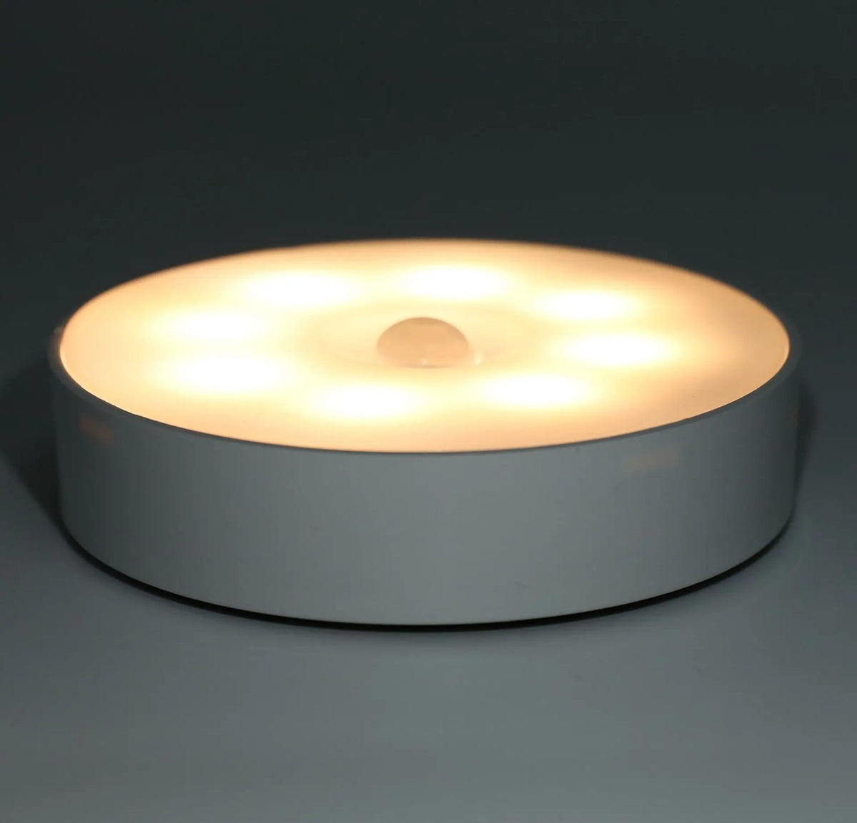 A round, white LED night light with a central sensor is shown on a dark surface.