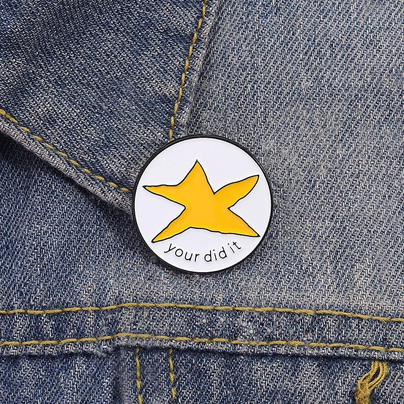 A yellow star enamel pin with the text your did it on a denim fabric background.