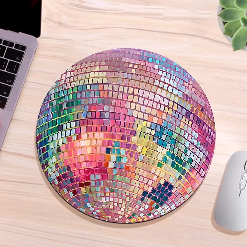 A pink disco ball mouse pad with a non-slip rubber base resting on a wooden desk next to a computer mouse.