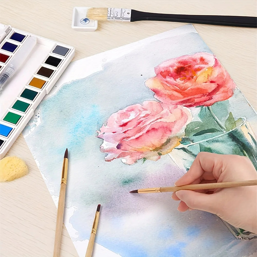A hand paints a pink rose watercolor on paper, surrounded by paint palettes and brushes.