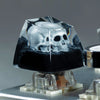 A monochrome skull resin mechanical keycap displayed on a clear stand.