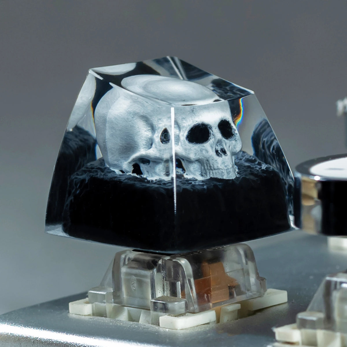 A monochrome skull resin mechanical keycap displayed on a clear stand.