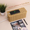 A black cable management box with a lid resting on a woven rattan box next to a potted plant.