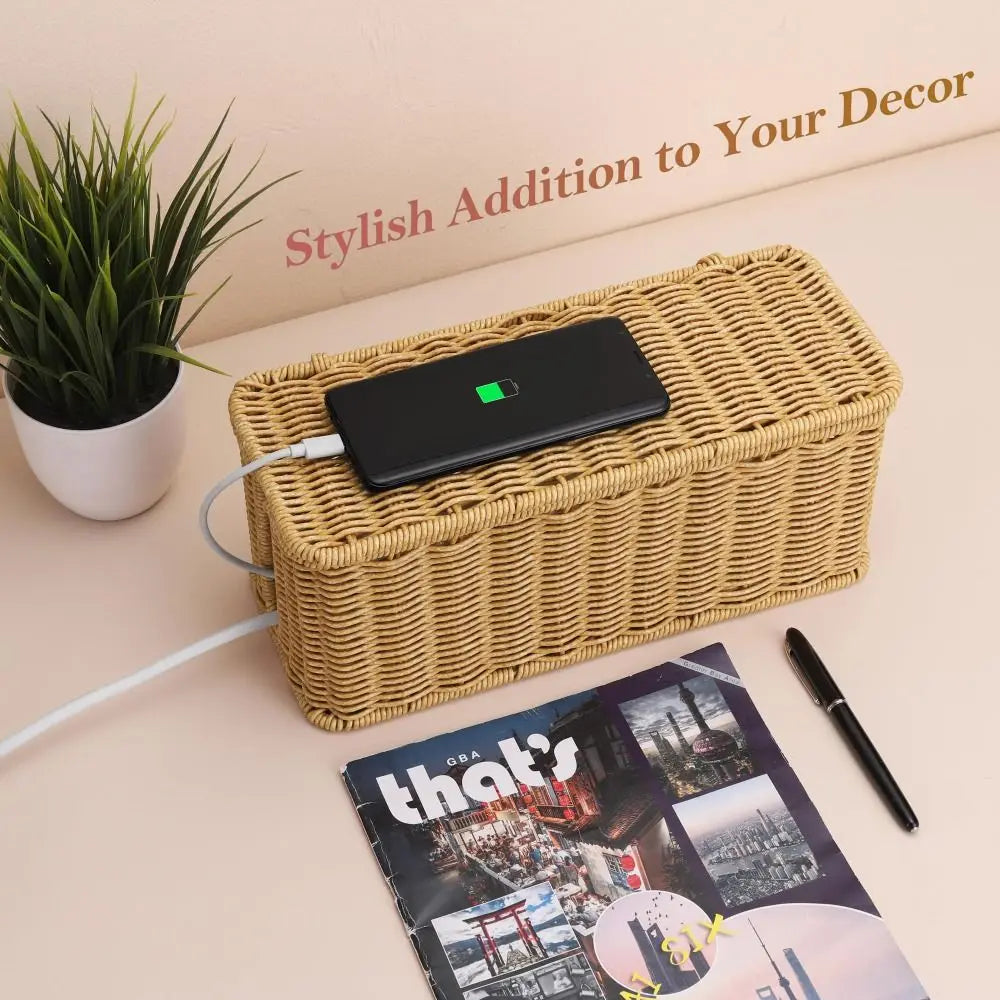 A black cable management box with a lid resting on a woven rattan box next to a potted plant.