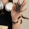 A hand holds a glossy tortoiseshell patterned protective case for AirPods next to a pair of white wireless headphones.
