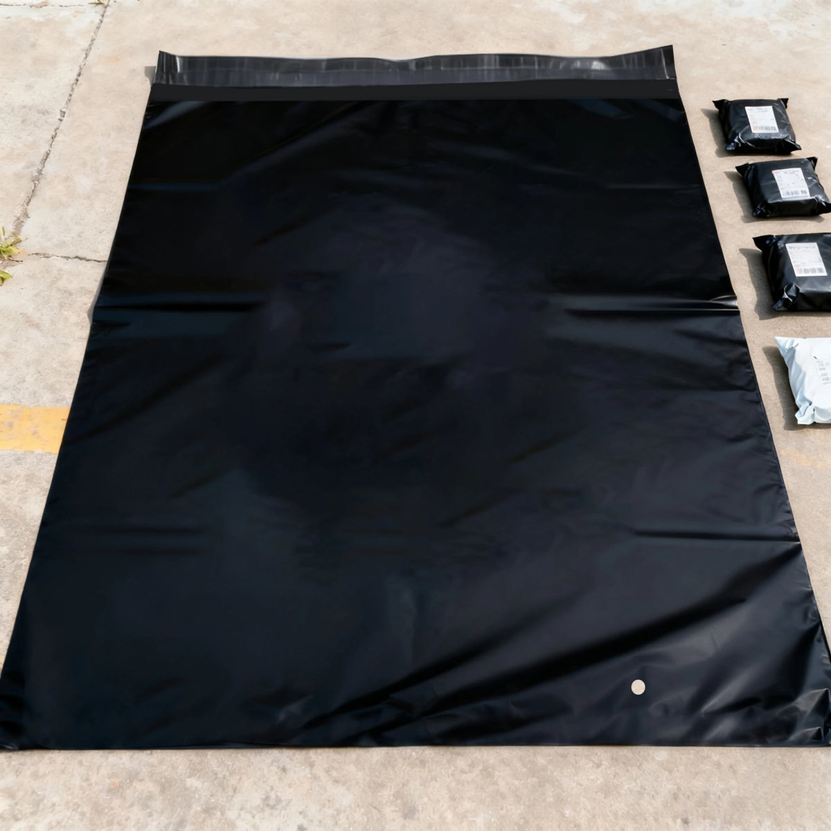 A black waterproof poly mailer is laid flat on a concrete surface with several smaller black mailers nearby.