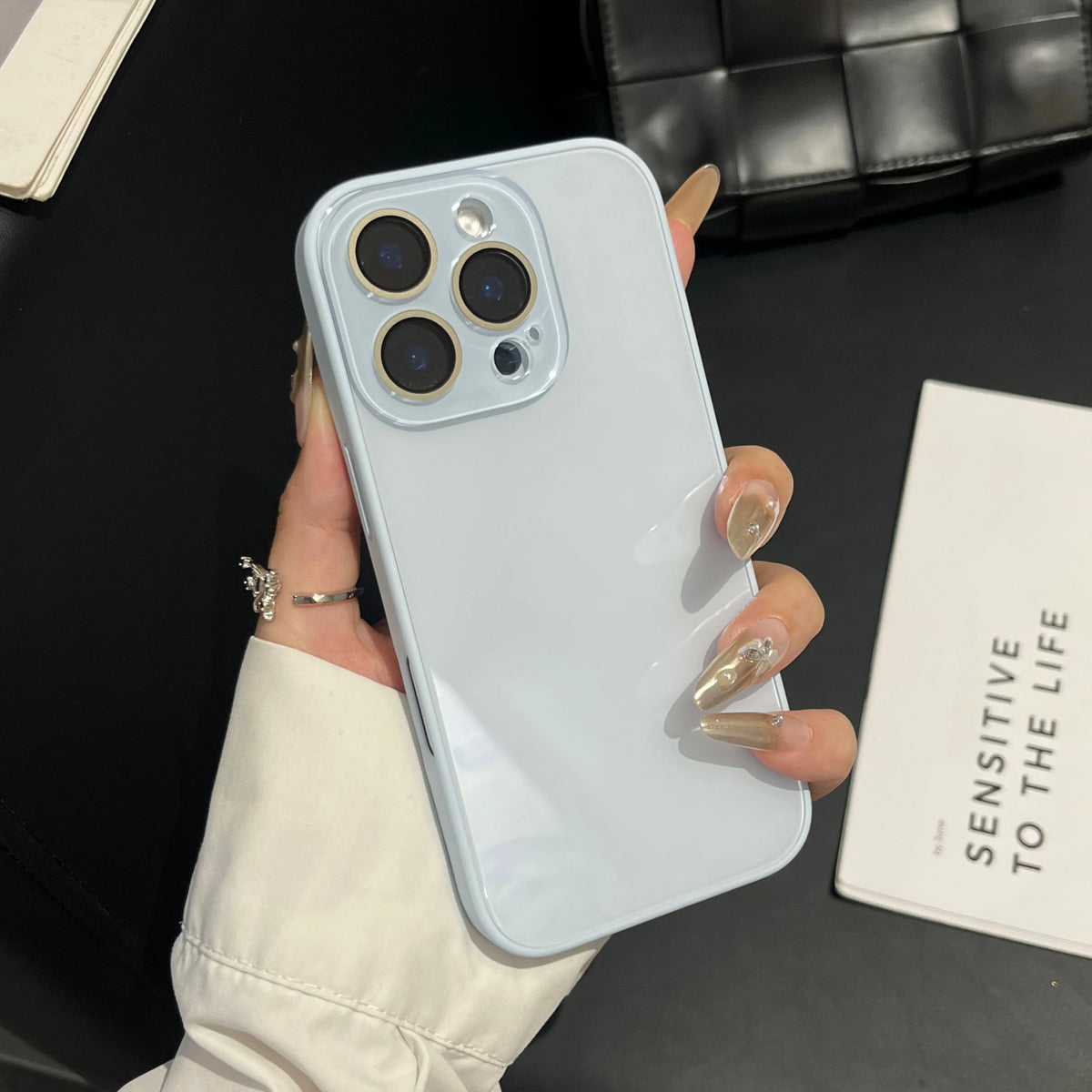 A hand holds a light blue iPhone case with a clear back, displaying the camera module and a gold nail polish.