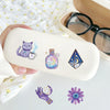 A light beige silicone case featuring purple magic witch stickers, including a cat, a potion bottle, and a geometric crystal.