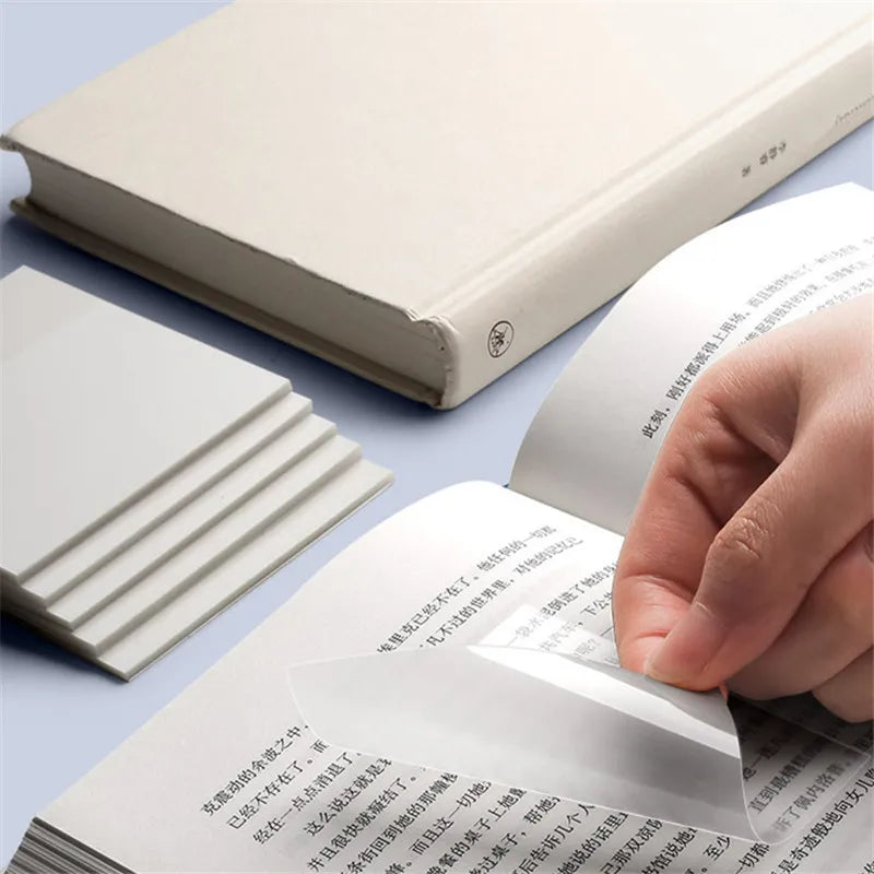A hand is holding and turning a sheet of transparent sticky notes memo pads next to a stack of white memo pads.
