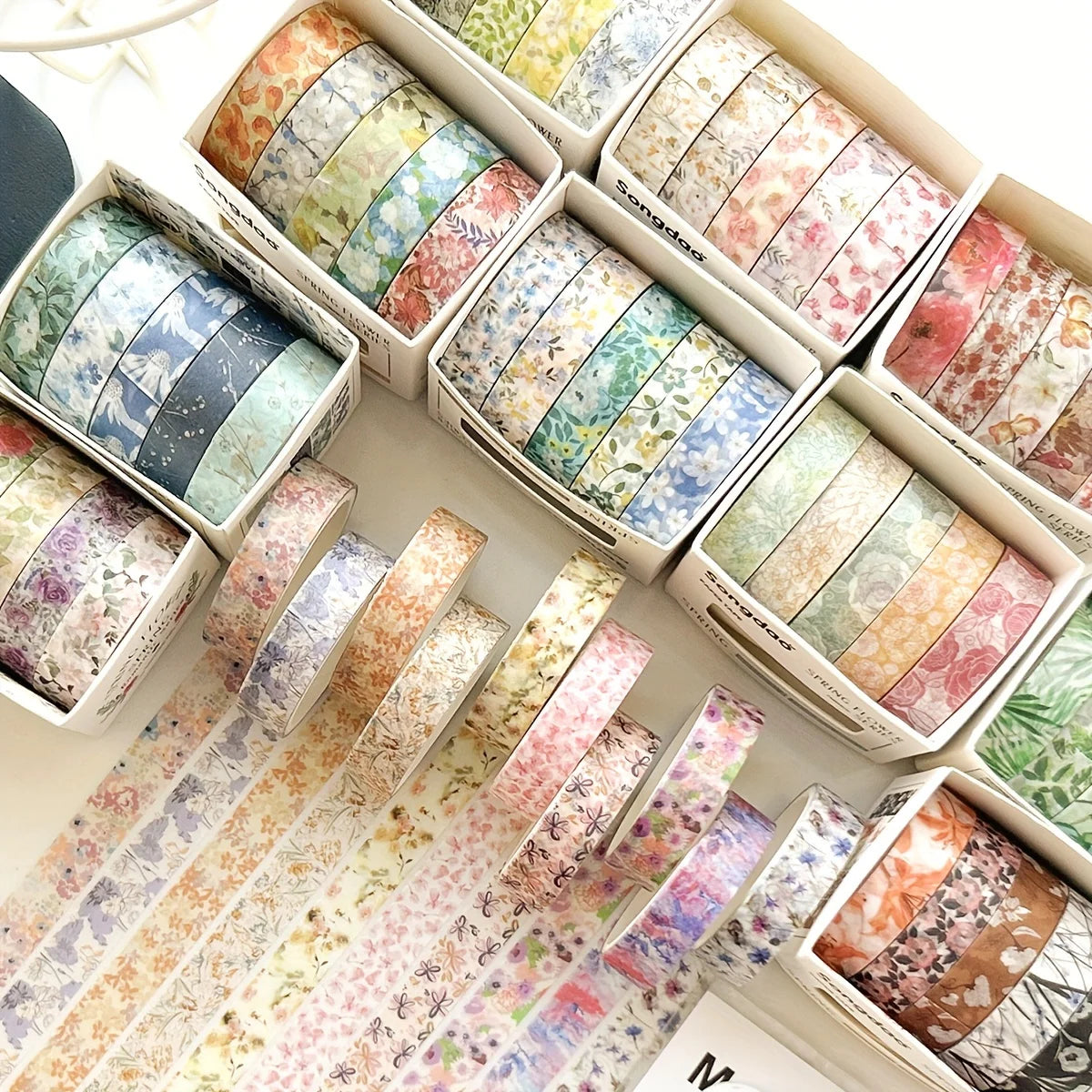 A collection of five rolls of floral washi tape in various patterns and colors displayed in cardboard boxes.