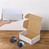 A hand is holding a white gift box and an open brown kraft cardboard box on a wooden surface.
