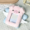A pink plush pig shaped photocard holder with white wings resting on a light blue fabric.