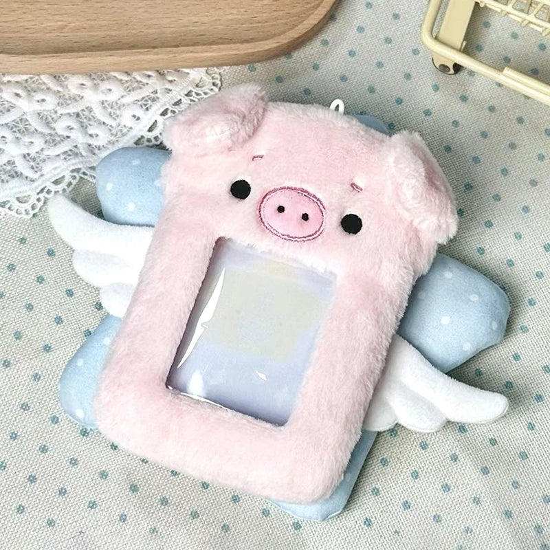 A pink plush pig shaped photocard holder with white wings resting on a light blue fabric.
