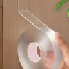 A hand is holding a roll of clear double-sided adhesive tape with a dispenser attached, against a wooden background.